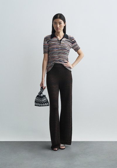 A nő áll, egyik kezét csípőre téve, színes, rövid ujjú kötött felsőt visel, áttetsző fekete mintás, bő szárú nadrágot, és egy fekete-fehér cikkcakk mintás táskát tart.