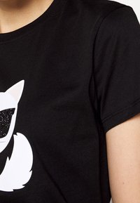 Camiseta de algodón negra con un gráfico de un gato de dibujos animados en blanco que lleva gafas de sol con purpurina negra, de mangas cortas y cuello redondo.