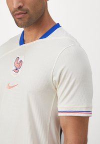 Maglia in jersey crema con texture a coste, colletto blu, accenti a strisce rosse e blu sulle maniche, con un emblema di gallo e il logo Nike.