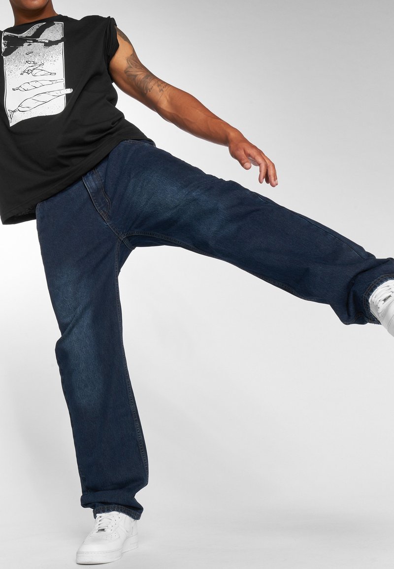 Zwarte mouwloze T-shirt met grafische print, donkerblauwe comfortabele jeans, witte sneakers. De jeans hebben een lichte vervaging en zichtbare naden.