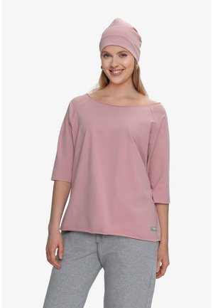 Uśmiechnięta kobieta w luźnej różowej bluzce z rękawami do łokci, pasującej różowej czapce oraz jasnoszarych spodniach dresowych, stojąca na białym tle.