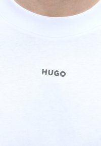 Valge ümara kaelusega t-särk väikese musta kirjaga "HUGO" kaeluse all, palja naha taustal.