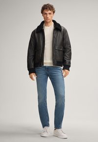 Giovane uomo con giacca di pelle nera con colletto di pelliccia, maglione bianco, jeans blu e sneakers bianche, in piedi su uno sfondo neutro.