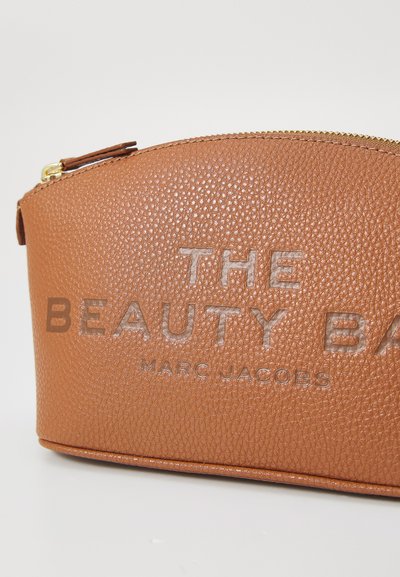 Bolso de cosméticos de cuero marrón texturizado con forma redondeada, que presenta texto en relieve: "EL BOLSO DE BELLEZA" y "MARC JACOBS" en beige claro. Detalles de cremallera dorada.