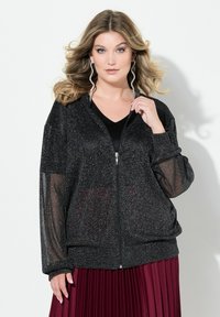 Giacca nera con zip, caratterizzata da una texture glitterata e maniche trasparenti, abbinata a un top nero a V e a una gonna plissettata bordeaux.