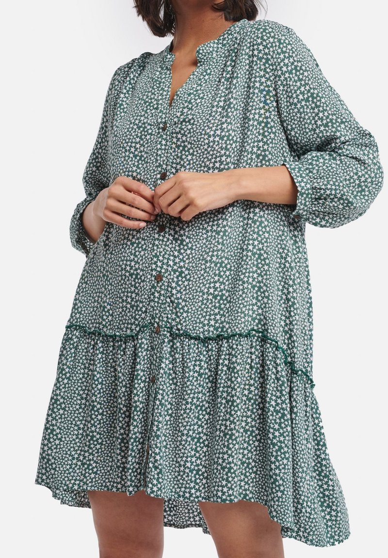 Femme portant une robe verte à manches longues boutonnée avec des motifs d'étoiles blanches, ajustant les boutons au niveau de la poitrine.