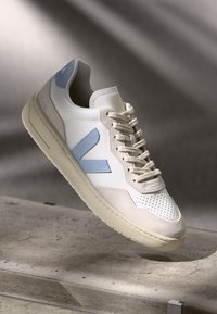 Hvit sneaker med lyseblå detaljer, teksturert lærpanel, rund tå og perforerte detaljer, med et klassisk snøringsdesign.