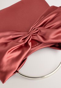 Rdeča satenasta torbica z okrasno pentljo in srebrnim okroglim ročajem. Gladek tekstura, pravokotna oblika in natančni šivi ob robovih.