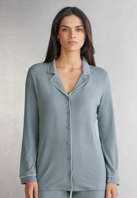 Lichtblauw pyjamajack van zachte stof, met een geknipt kraagje, lange mouwen, witte biezen en knopen aan de voorkant.