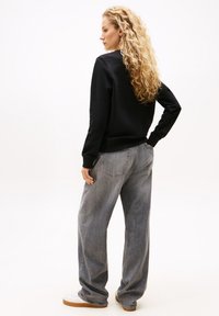 Felpa nera con collo a giro, abbinata a jeans grigi a vita alta e a taglio largo. Il modello ha i capelli ricci e indossa scarpe bianche con suole in gomma.