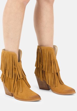 Stivaletti alla caviglia in camoscio color cuoio con frange a strati e tacchi impilati in legno indossati su gambe nude su sfondo bianco.