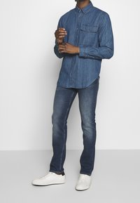 Camicia in denim con chiusura a bottoni e taschino sul petto, abbinata a jeans blu scuro e sneakers bianche. Il tessuto appare liscio con una finitura leggermente sbiadita.