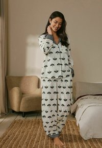 Weißes Pyjama-Set mit schwarzen Baum- und Regenschirmmustern. Enthält ein Hemd mit Kragen und eine Hose mit elastischem Bund; weicher Stoff mit kontrastierenden Details.