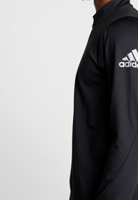 Schwarzes, athletic Langarmshirt mit hohem Kragen, mit weißem Adidas-Logo auf der linken Schulter. Glatte Textur, enganliegendes Design.