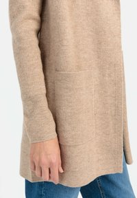 Beige Rippstrick-Cardigan mit langen Ärmeln, seitlichen Schlitzen und vorderen Taschen. Weiche Textur und mittellanges Design. Getragen über mittelblauen Jeans.