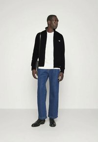 Černá bunda na zip přes bílé tričko, modré široké džíny a černé kotníkové boty. Jednoduchý design s logem na bundě.