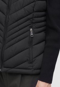 Gros plan d’une veste matelassée noire avec des coutures diagonales et une poche zippée à côté du bras d’une personne portant une manche noire.