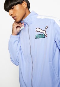 Lys blå Puma vindjakke med høj krave, lynlås, broderet logo og hvide skulderakcenter. Glat tekstur.