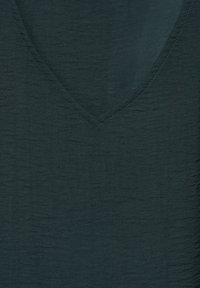 Mörkgrön, texturerad tyg med V-ringning. Ytan har ett subtilt, ojämnt mönster och kontrasterande sömmar runt halsringningen.