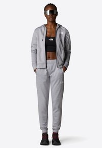 Grijze rits hoodie met trekkoord en logo; zwarte sport-bh; grijze joggingbroek; zwarte schoenen met rode accenten; zonnebril.