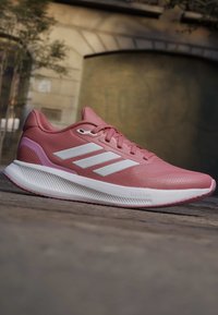 Chaussure de course Adidas rose avec des bandes blanches et une semelle blanche épaisse portant l'étiquette "Cloudfoam" sur un fond de pavé extérieur.