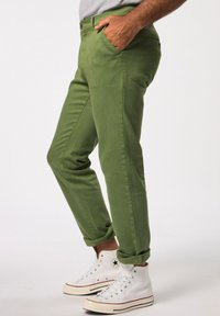 Chino in cotone verde con tasche laterali, taglio slim e orli arrotolati, abbinati a sneakers bianche alte. Dettaglio di chiusura con bottone semplice.