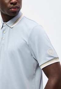 Άνδρας που φοράει ανοιχτό μπλε κοντομάνικο πόλο με γιακά και άκρες μανικιών με μπεζ και λευκές ρίγες, με λογότυπο στο πάνω μέρος του μπράτσου.