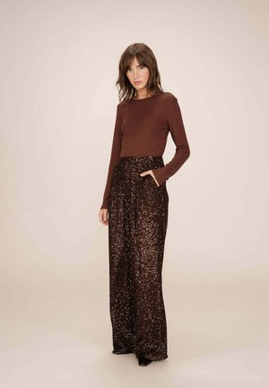 Femme debout portant un haut marron à manches longues et un pantalon pailleté marron foncé taille haute et large, avec une main dans la poche.