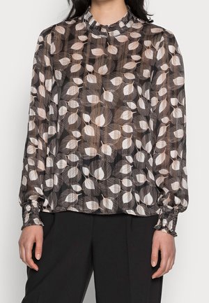 Femme portant un chemisier noir transparent avec des motifs abstraits de feuilles beige et blanc et des poignets froncés, associé à un pantalon noir.