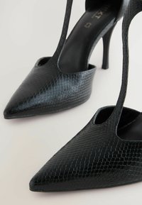 Zwarte pointed-toe hoge hakken met een textuur van een reptielpatroon en uitsnijdingen aan de zijkant, met dunne stiletto-hakken en een glad interieur.