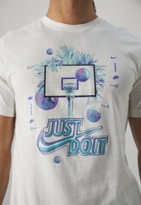 Camiseta de algodón blanca con un gráfico de un aro de baloncesto rodeado de palmeras y balones de baloncesto flotantes, acentuado con el texto "JUST DO IT" en multicolor.