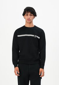 Young man with dark curly hair wearing a black sweatshirt with white horizontal stripes and black pants, standing against a white background.