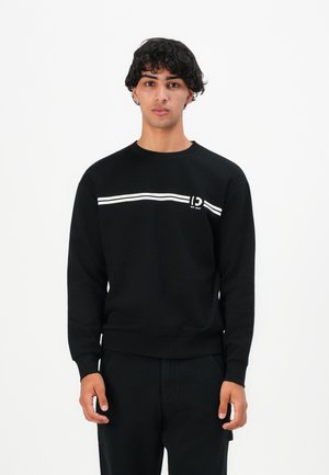 Young man with dark curly hair wearing a black sweatshirt with white horizontal stripes and black pants, standing against a white background.