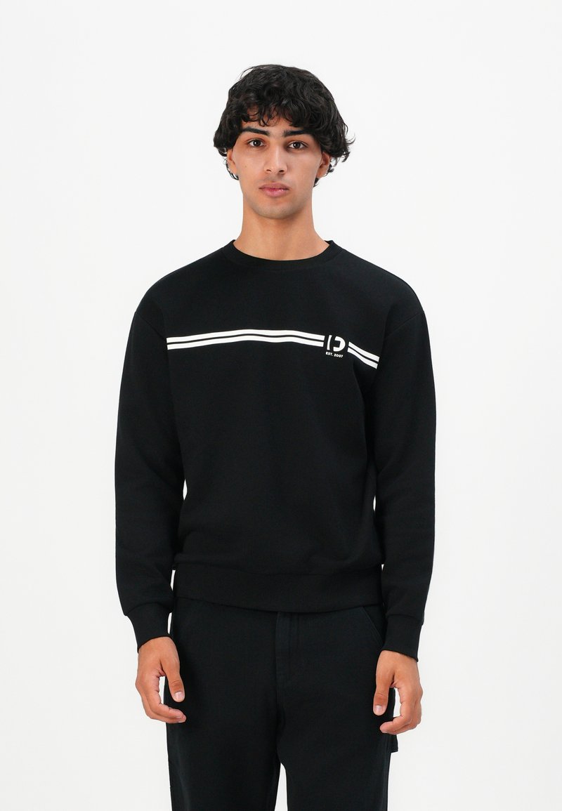 Young man with dark curly hair wearing a black sweatshirt with white horizontal stripes and black pants, standing against a white background.