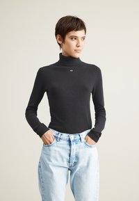 Maglione a collo alto nero a coste con maniche lunghe, dotato di un piccolo logo, abbinato a jeans di denim azzurri a vita alta.