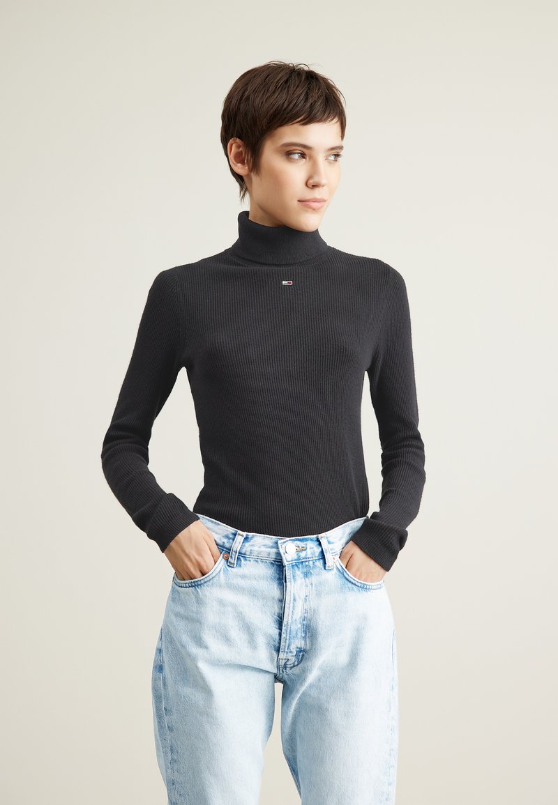 Maglione a collo alto nero a coste con maniche lunghe, dotato di un piccolo logo, abbinato a jeans di denim azzurri a vita alta.