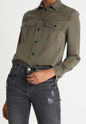 Olivegrünes Hemd mit Knöpfen, zwei Brusttaschen, langärmlig bis zu den Ellenbogen hochgerollt, kombiniert mit schwarzen, abgewetzten Jeans.