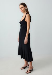 Frau in einem schwarzen ärmellosen Midikleid mit Rüschendetails, trägt blumige Ohrringe und schwarze Absatzsandalen, steht vor einer einfarbigen Wand.