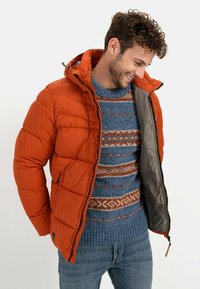 Puffed orange jacket with a hood, zip closure, and inner gray lining, worn over a patterned blue sweater featuring orange and cream details.