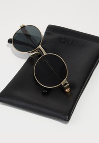 Gafas de sol redondas negras con montura de metal dorado, reposando sobre un estuche de cuero negro con la palabra "QUAY" grabada.