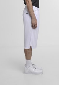 Personne portant un short blanc en maille arrivant aux genoux, des chaussettes blanches montantes et des baskets Nike blanches, debout de profil sur un fond clair uni.