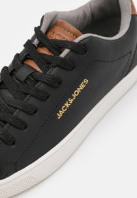 Baskets en cuir noir avec lacets noirs, semelle blanche, doublure intérieure grise, languette marron et logo doré "JACK&JONES" sur le côté.