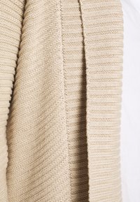 Beiger gestrickter Pullover mit verschiedenen Rippen- und strukturierten Strickmustern, die im Stoff nahe aufgenommen sichtbar sind.