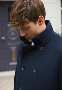 Marineblauer Wollmantel mit stehendem Kragen, doppelreihigem Design und schwarzen Knöpfen. Strukturierter Stoff und taillierte Passform.