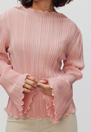 Woman wearing a pink pleated long-sleeve top with ruffled cuffs and hem, hands loosely clasped in front.
