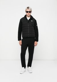 Sort puffervest med fleeceærmer, lynlås foran og logo-patch; parret med sorte bukser og hvide sneakers. Model iført solbriller.