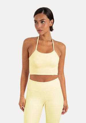 Femme portant un crop top halter jaune pâle et des leggings taille haute assortis, regardant sur le côté sur un fond uni.