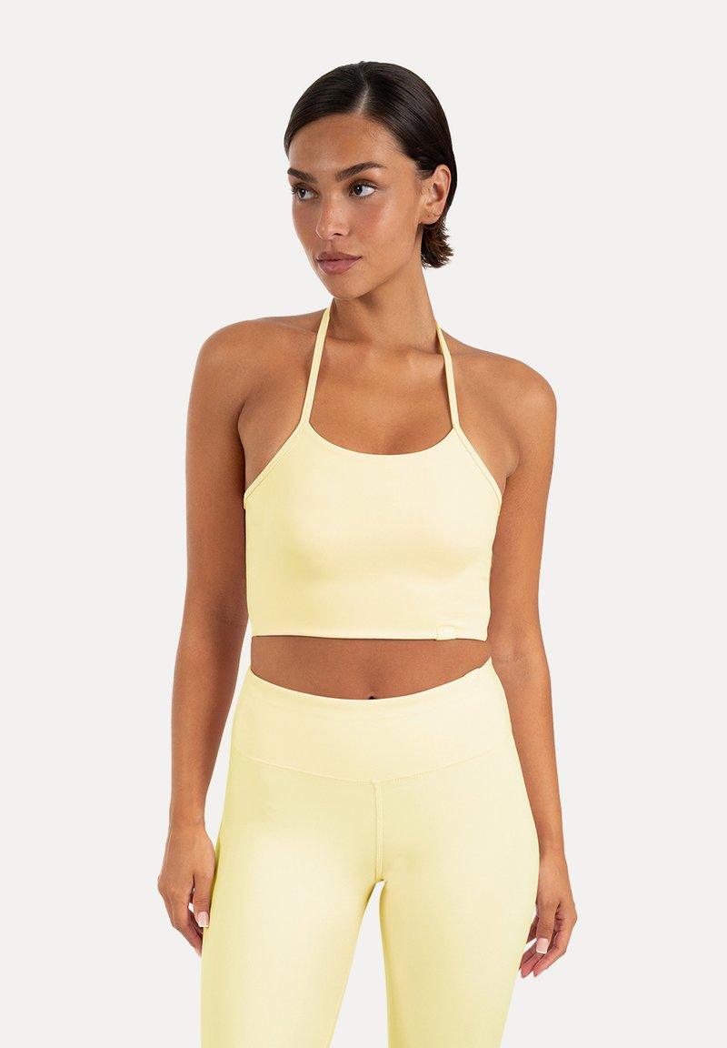 Femme portant un crop top halter jaune pâle et des leggings taille haute assortis, regardant sur le côté sur un fond uni.