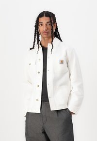 White denim jacket with gold snap buttons, front pockets, and a logo patch. Worn over a dark gray shirt and paired with gray pants.