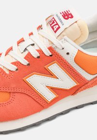 New Balance 574 sneakers feature orange suede and mesh, white leather accents, a thick white midsole, and visible branding on the tongue.